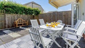 Un patio con una mesa dispuesta para cuatro personas, rodeado de sillas blancas, un banco de madera y un toldo amarillo a rayas.