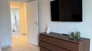 A modern living room with a flat-screen TV mounted on the wall, a wooden cabinet with drawers, and a patterned rug on the floor. - The Bacchae - Urban Apartment (Siracusa)