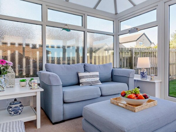 A conservatory with a grey sofa, a small table with a lamp, and a bowl of fruit on a tray. - 3 Bed in Seahouses (oc-cn064) (Seahouses)