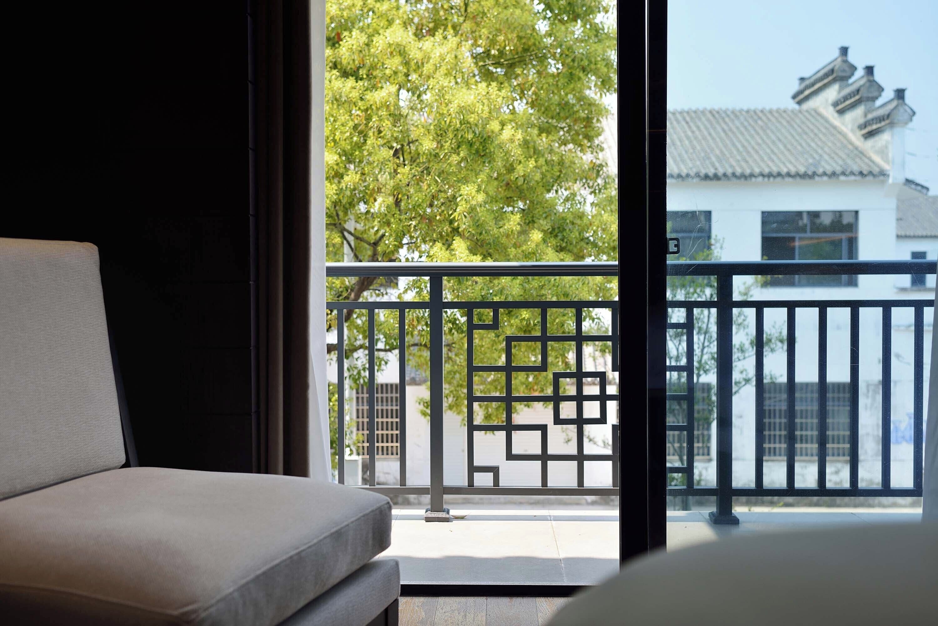 A room with a sofa, a balcony with a metal railing, and a view of trees and a building.