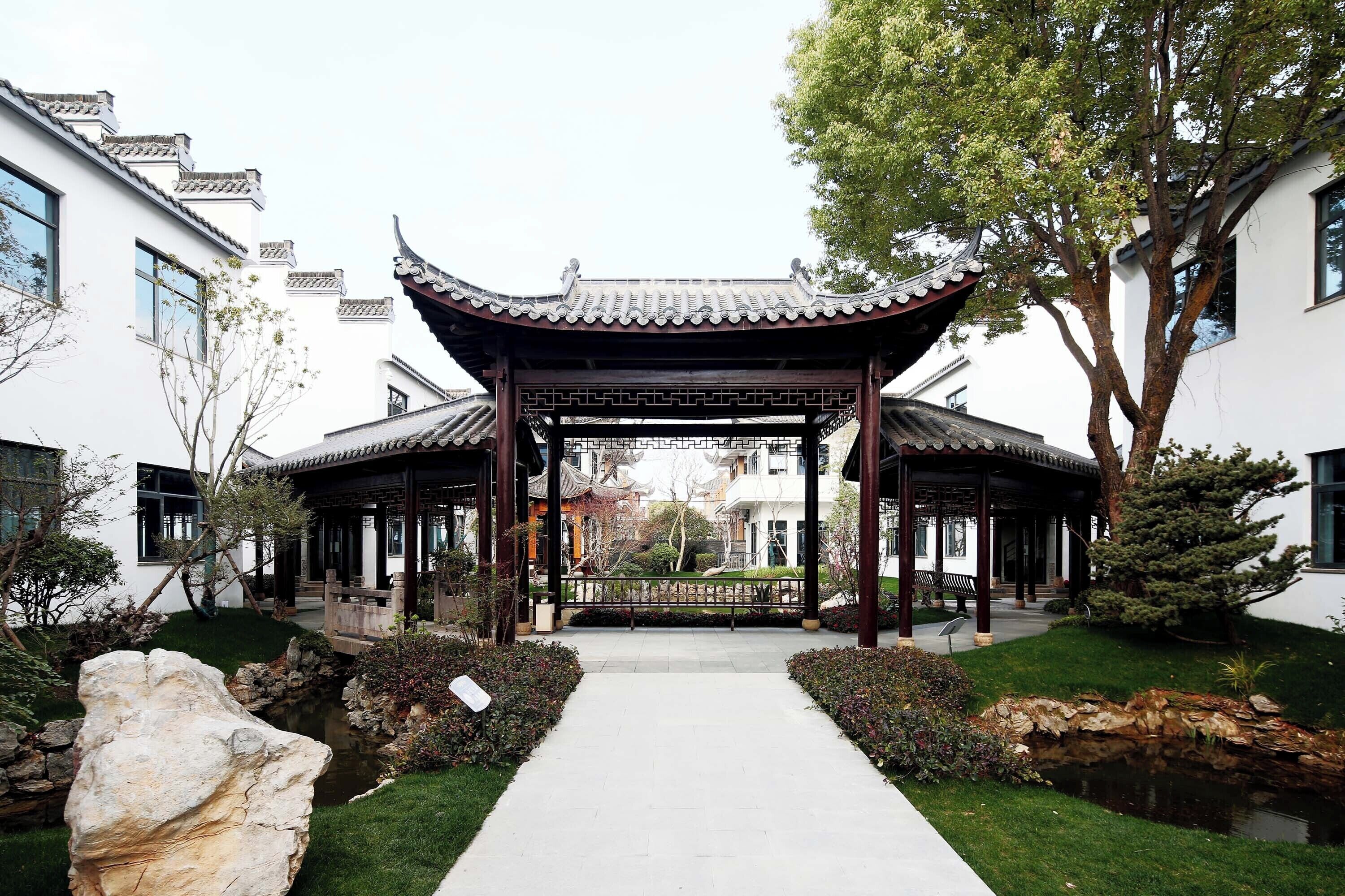 A traditional Chinese pavilion with a tiled roof, surrounded by a well-maintained garden with a pathway leading to it.