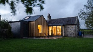 Une maison de campagne à l’aspect rustique, avec une façade en pierre, une extension en bois et un intérieur visible comprenant un coin repas.