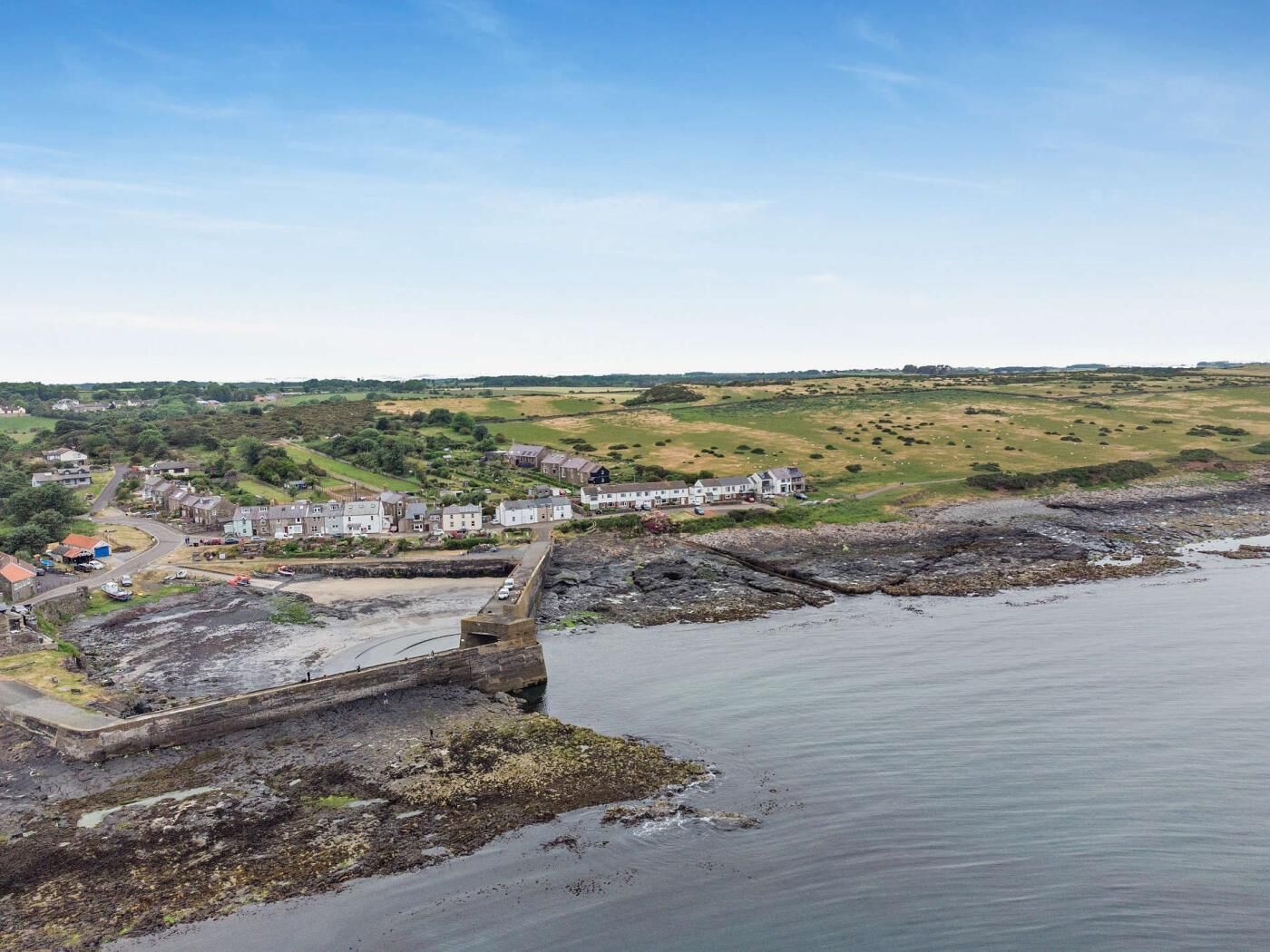 Een kustplaats met een opvallende stenen pier die het water in reikt, omringd door woonhuizen en groene velden.