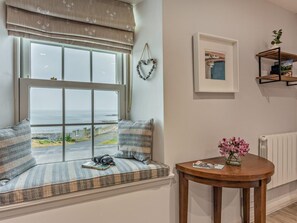 A room with a large window offering a view of the sea, a cozy seating area with cushions, a small table with a vase of flowers, and a framed picture on the wall. - 2 Bed in Alnwick (oc-cn096) (Dunstan)