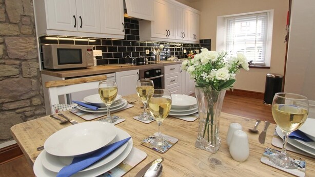 Una cocina moderna con armarios blancos, un microondas y una mesa de comedor preparada para cuatro, con platos y copas de vino blanco.