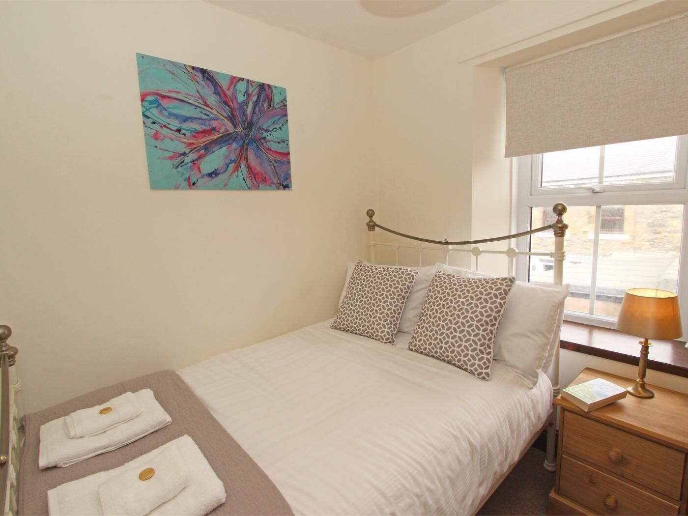 Un dormitorio con cama, almohadas, una mesita de noche, lámpara, ventana con vistas y un cuadro en la pared.