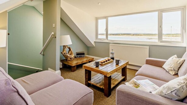A living room with a view of the beach, a staircase, and a table with a lamp and a radio. - 3 Bed in Beadnell (oc-cn119) (Beadnell)