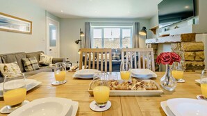 A dining area with a wooden table set for breakfast, a sofa, and a TV mounted on the wall. - 3 Bed in Beadnell (oc-cn119) (Beadnell)