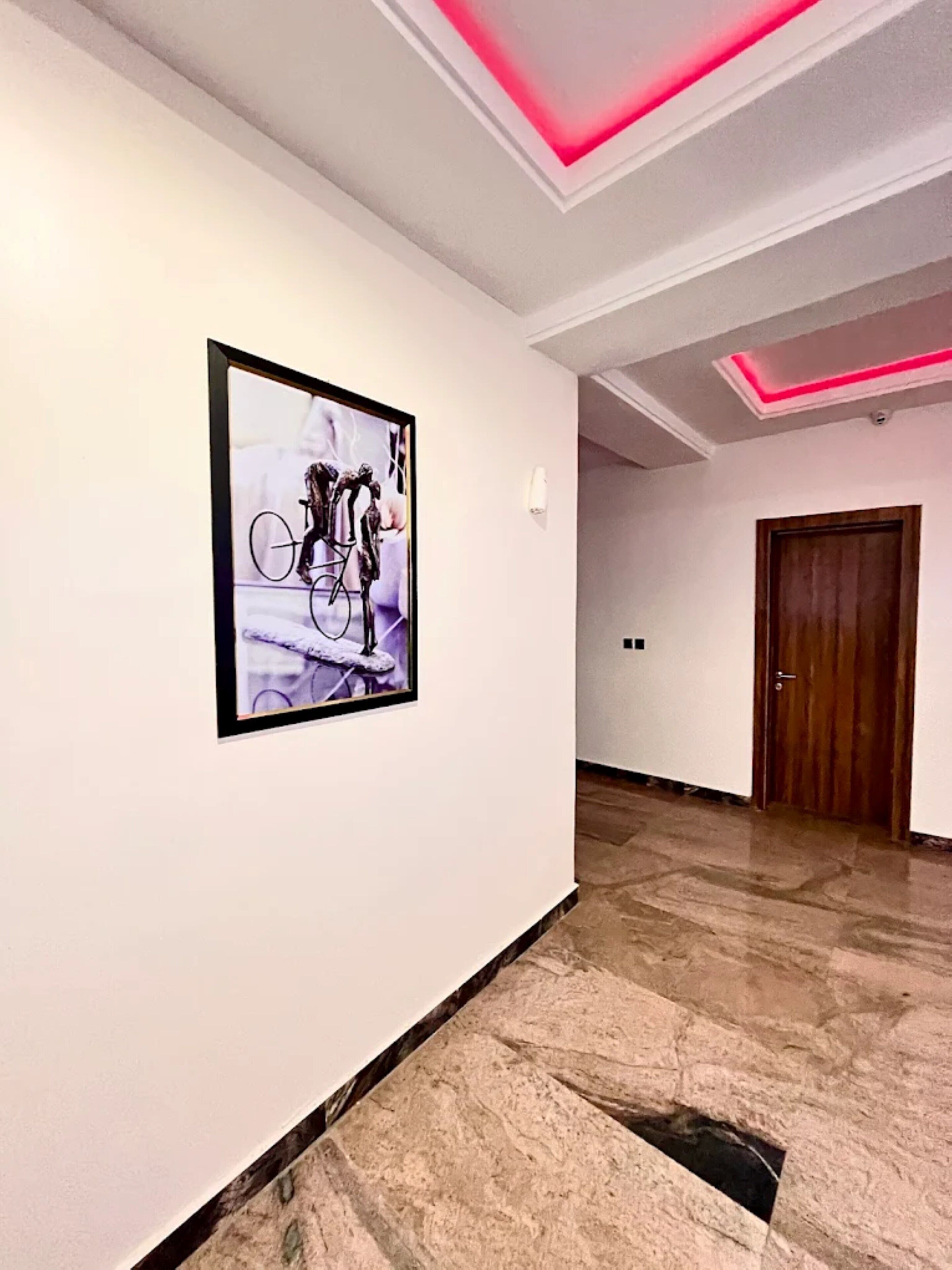 A hallway with a framed abstract artwork, a wooden door, and a ceiling with pink lighting.
