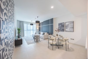 A modern living room with a sofa, dining table, and chairs, a large window with curtains, and a patterned wall. - Bloomfields 1BR in Radiant Viewz 1  (Abu Dhabi)
