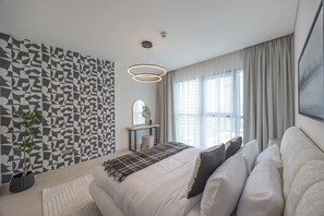 A modern bedroom with a geometric-patterned wall, a bed with white bedding, a round pendant light, and a large window with curtains. - Bloomfields 1BR in Radiant Viewz 1  (Abu Dhabi)
