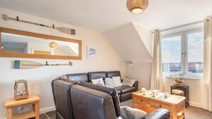 A modern living room with a sectional sofa, a small wooden table, a fireplace, and a large window with a view. - 3 Bed in Seahouses (oc-cn112) (Seahouses)