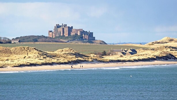 A coastal landscape with a castle on a hill, sandy beach, and calm sea. - 3 Bed in Seahouses (oc-cn112) (Seahouses)