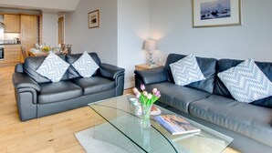 A modern living room with two black leather sofas, a glass coffee table, and a dining area in the background. - 3 Bed in Seahouses (oc-cn120) (Seahouses)