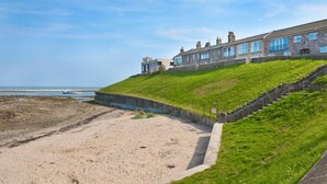 A coastal building with a stone facade, a grassy slope, and a sandy beach area. - 3 Bed in Seahouses (oc-cn120) (Seahouses)