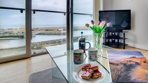 A modern living room with a large glass door leading to a view of the sea, a coffee maker, tulips in a vase, a TV, and a magazine on the table. - 3 Bed in Seahouses (oc-cn120) (Seahouses)