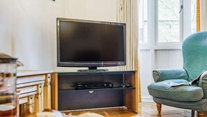 Un salon doté d’un téléviseur à écran plat, d’un meuble de rangement en bois, d’un fauteuil vert et d’une fenêtre avec des rideaux.