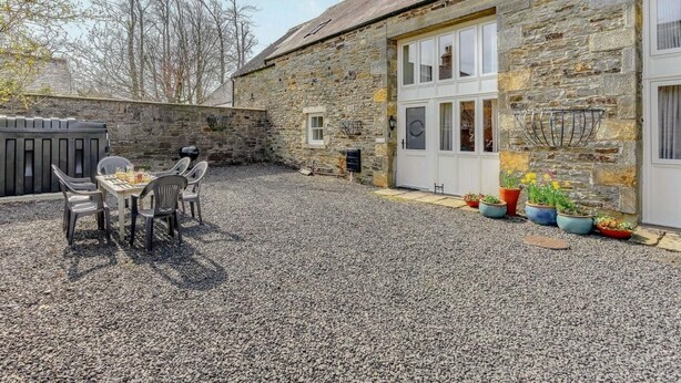 Un bâtiment en pierre avec une cour recouverte de gravier, une table à manger avec des chaises et des plantes en pot.