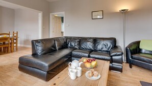 Ein Wohnzimmer mit einer Eckcouch, einem Couchtisch mit Tassen und Gebäck, einem Essbereich mit Stühlen und einer Stehlampe.