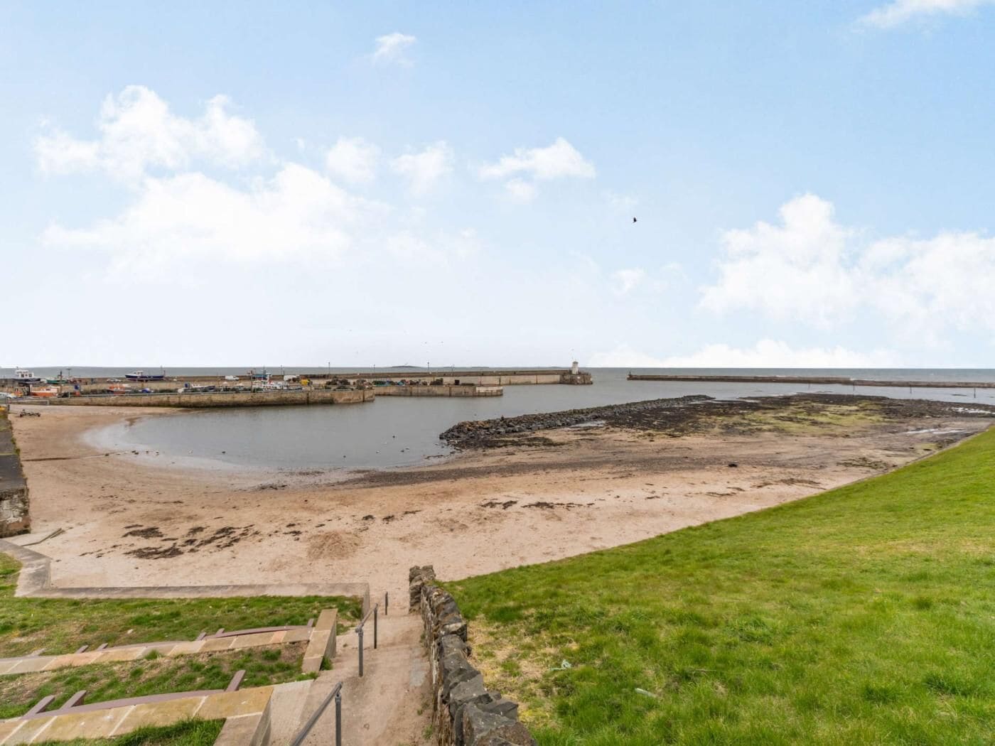 A coastal landscape with a sandy beach, a grassy area, and a stone wall.