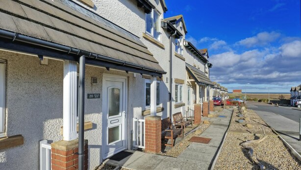 A row of terraced houses with a clear sky, a visible number '20' on the front door, and a gravel path leading to the entrance. - 2 Bed in Seahouses (oc-cn135) (Seahouses)