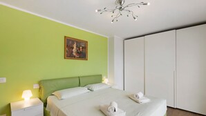 A bedroom with a green wall, a bed with white bedding, a painting, a bedside lamp, and a wardrobe.