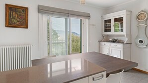 A kitchen with a large window, a radiator, a hanging light fixture, a cabinet with glass doors, and a wall-mounted clock.