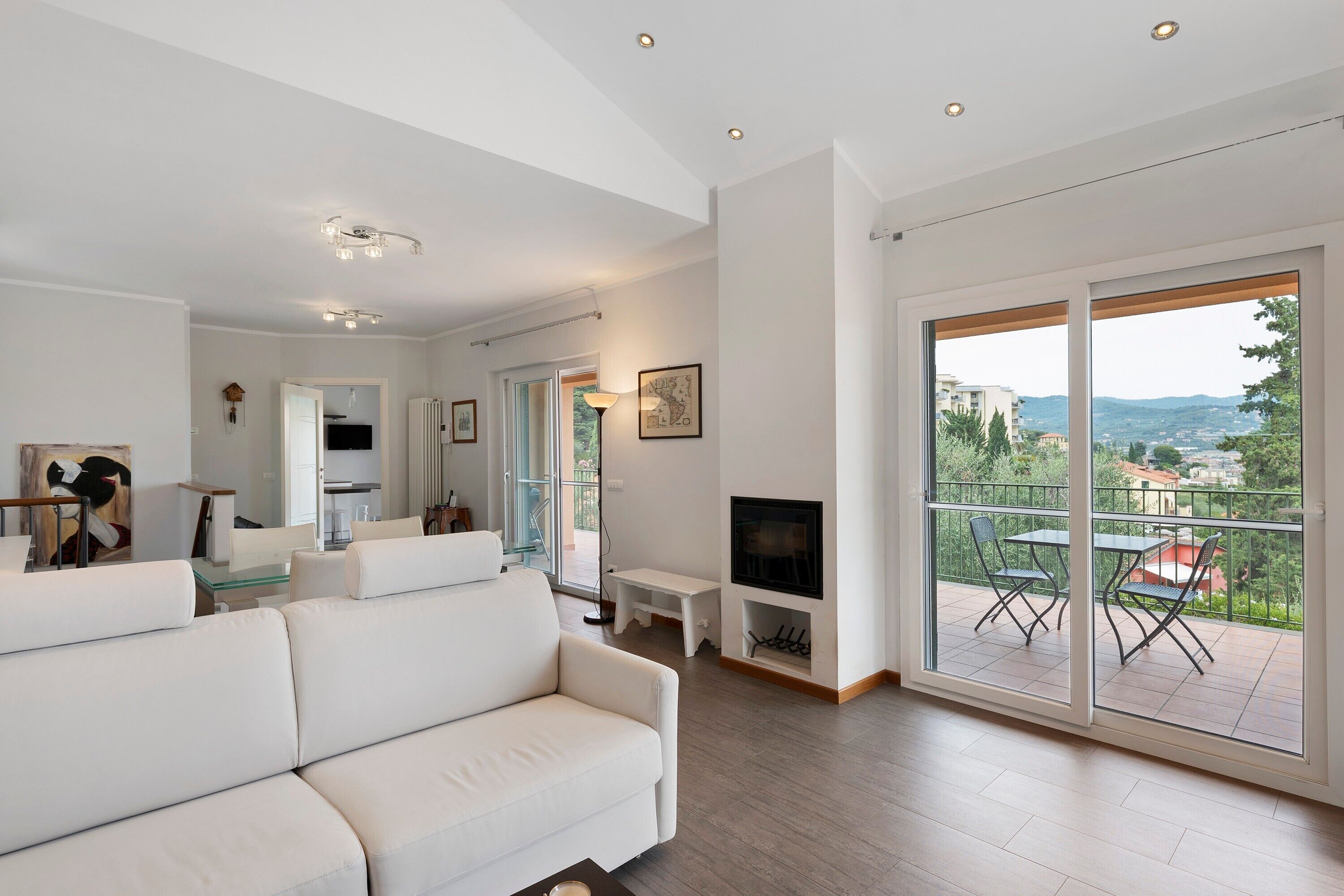 A modern living room with a white sofa, a flat-screen TV, and a balcony with a view of the city.