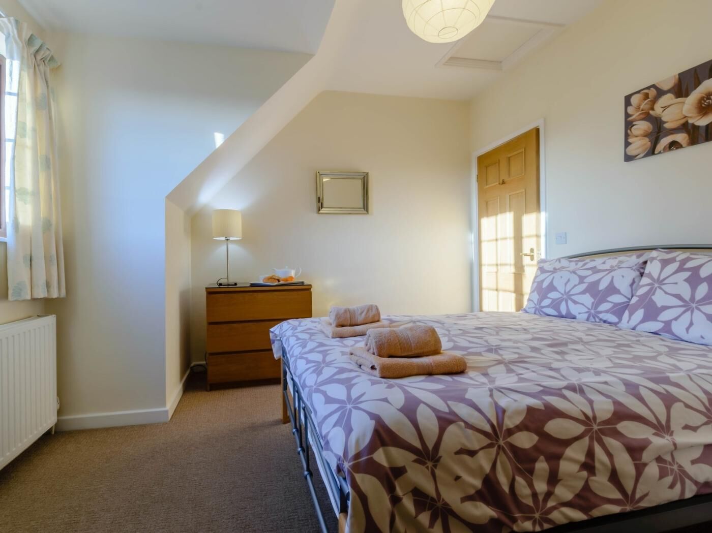 A bedroom with a bed, a nightstand, a lamp, a dresser, and a window with curtains.