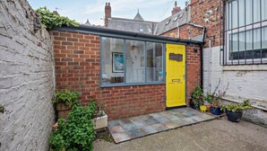 A brick house with a yellow door, a window, and a small garden area with potted plants. - 5 Bed in Tynemouth (oc-cn125) (Tynemouth)