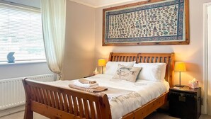 Uma cama de madeira com roupa de cama branca, duas luminárias de cabeceira, um criado-mudo e uma tapeçaria grande na parede.