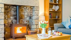 A cozy room with a stone fireplace, a wooden table with magazines and a vase of tulips, and a bookshelf with decorative items. - 3 Bed in Seahouses (oc-cn103) (Seahouses)