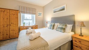 A bedroom with a bed, a wooden wardrobe, a window with curtains, a bedside table with a lamp, and a painting on the wall. - 3 Bed in Seahouses (oc-cn103) (Seahouses)