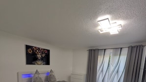 A modern bedroom with a bed, bedside tables, a rug, a painting, and a window with curtains. - Vacation rental, comfortable and modern, designed for discerning guests, near the main train station (Freudenstadt)