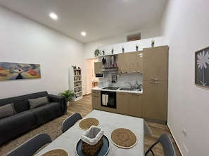 Een ruimtelijk ingerichte leefruimte met kitchenette, een eethoek met tafel en stoelen, een bank en een boekenkast.
