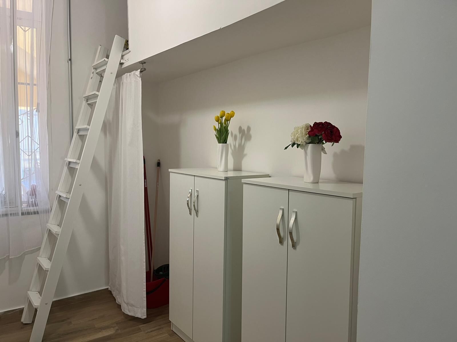 A white wardrobe with two doors, a white ladder leaning against the wall, and a white curtain partially drawn.