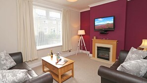 A living room with two leather sofas, a wooden coffee table, a fireplace, a television mounted on the wall, and a window with curtains. - 3 Bed in Seahouses (oc-cn182) (Seahouses)