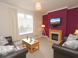 A living room with two leather sofas, a wooden coffee table, a fireplace, a television mounted on the wall, and a window with curtains. - 3 Bed in Seahouses (oc-cn182) (Seahouses)