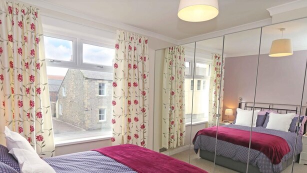 A bedroom with two beds, floral curtains, a large mirror, and a view of a stone building outside. - 3 Bed in Seahouses (oc-cn182) (Seahouses)