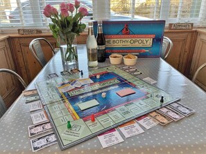 Ein Gesellschaftsspiel auf einem Tisch, mit einer Spielschachtel mit dem Titel 'The Bothy-Opoly', einer Flasche und einer Vase mit Blumen.
