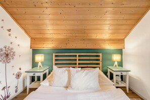 Ein Schlafzimmer mit Holzbalkendecke, einem grünen Kopfteil, zwei Nachttischen mit Lampen sowie einem Bett mit weißer Bettwäsche.