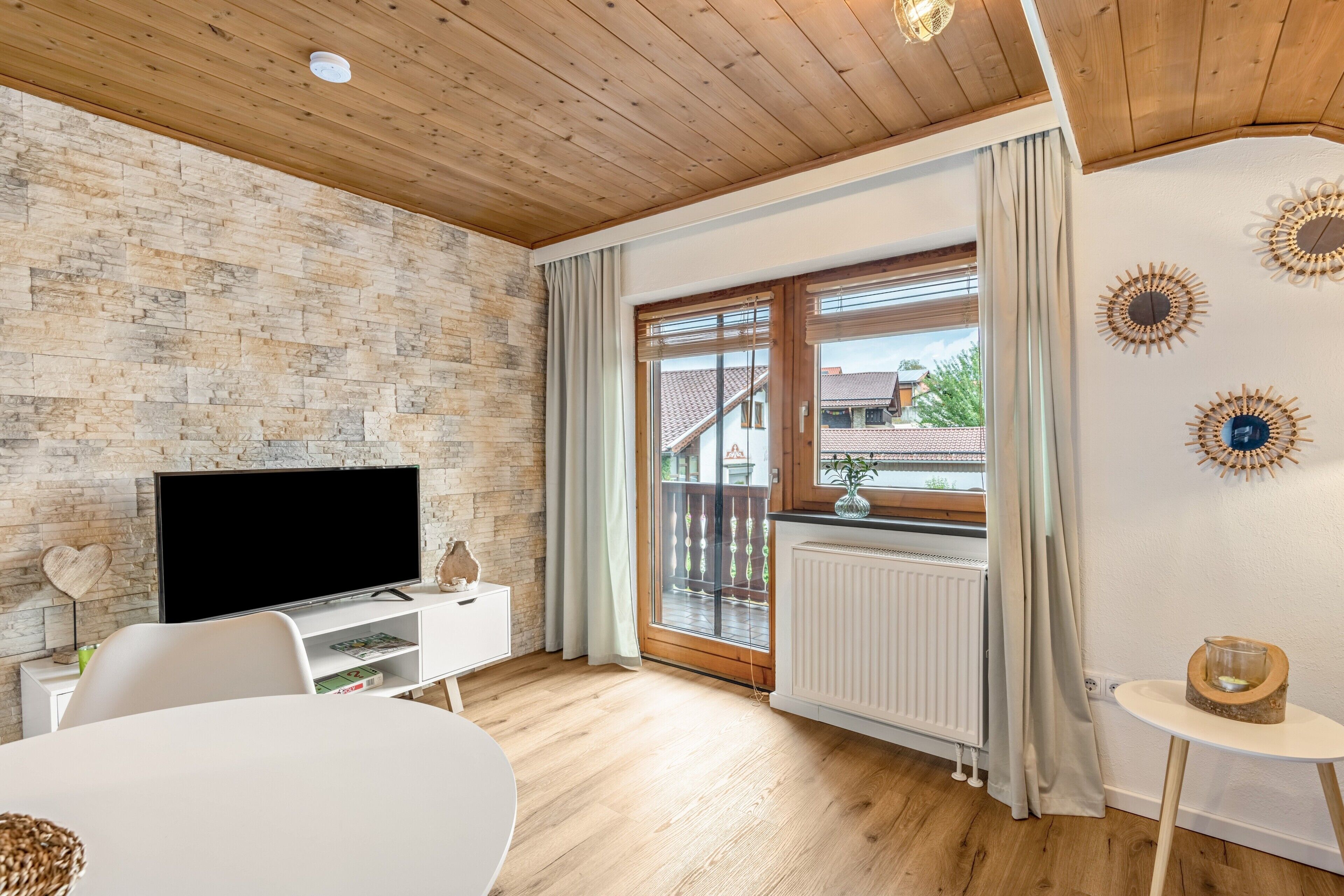 Ein modernes Wohnzimmer mit Steinwand, Holzboden, einem Flachbildfernseher, einem weißen Tisch und einem Balkon mit Blick auf Gebäude.