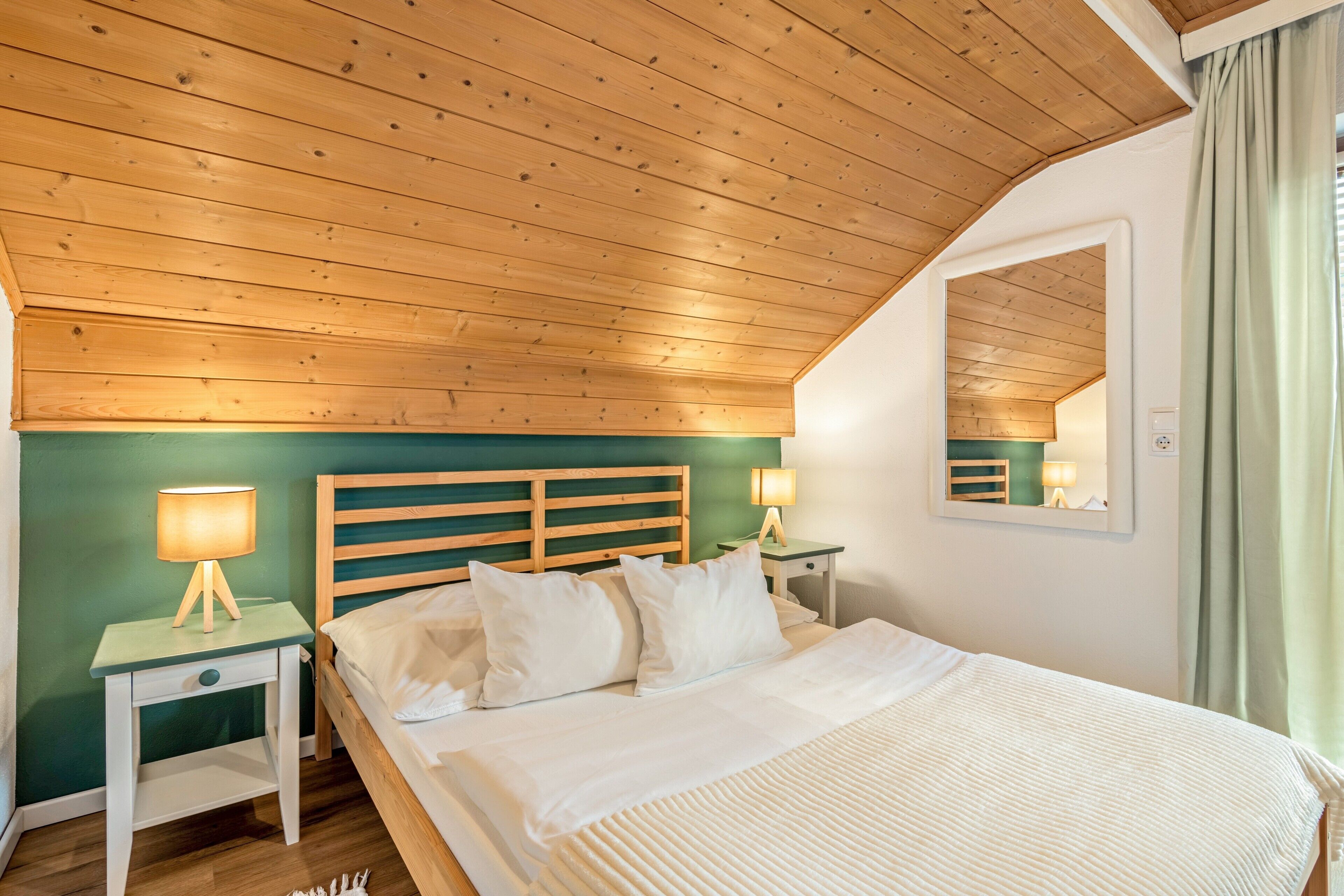 A bedroom with a wooden ceiling, a bed with white bedding, two bedside tables with lamps, and a mirror on the wall.