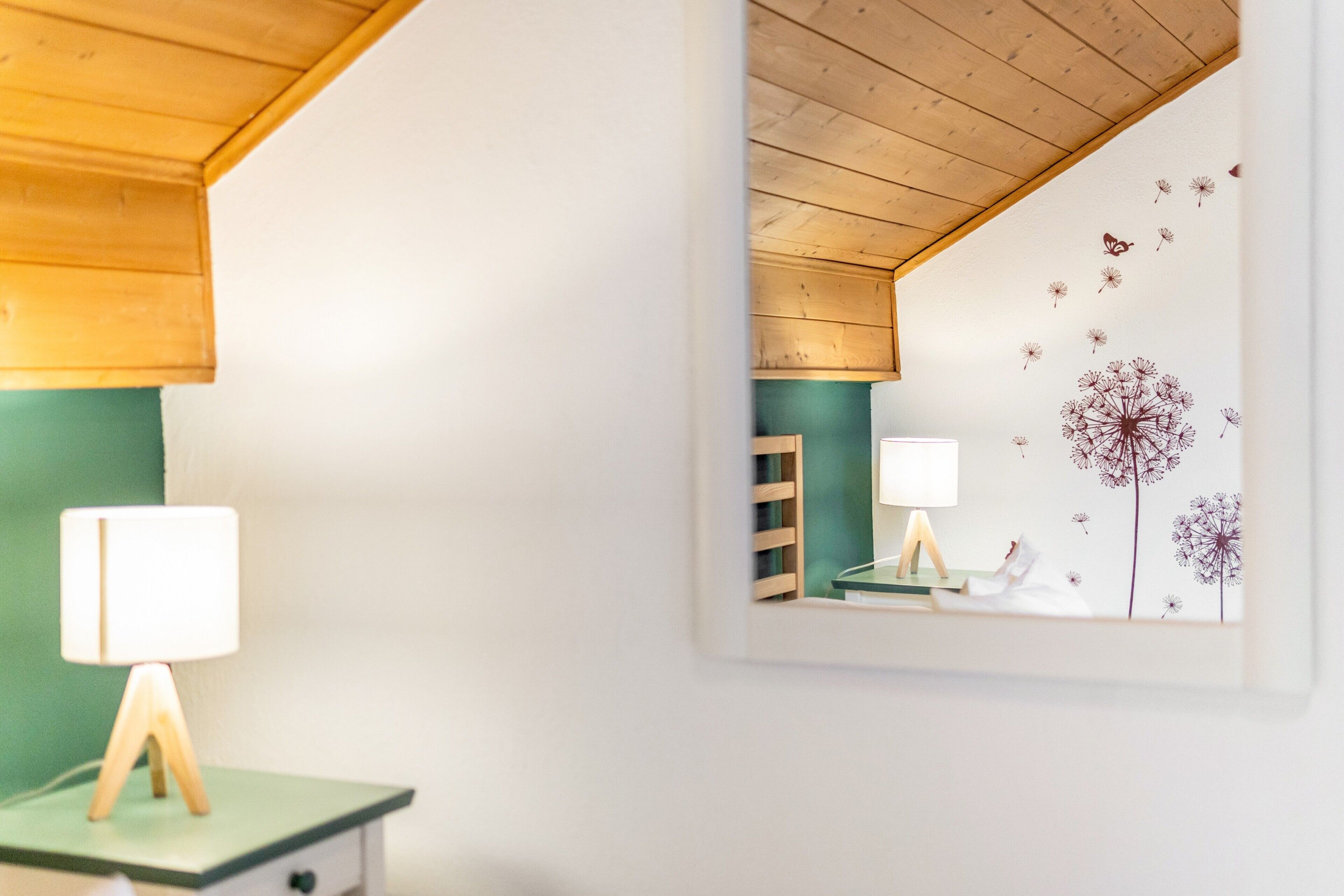 Ein Zimmer mit Holzboden, grüner Wand und Spiegel, der eine Lampe und eine Wand mit Löwenzahndeko zreflektiert.