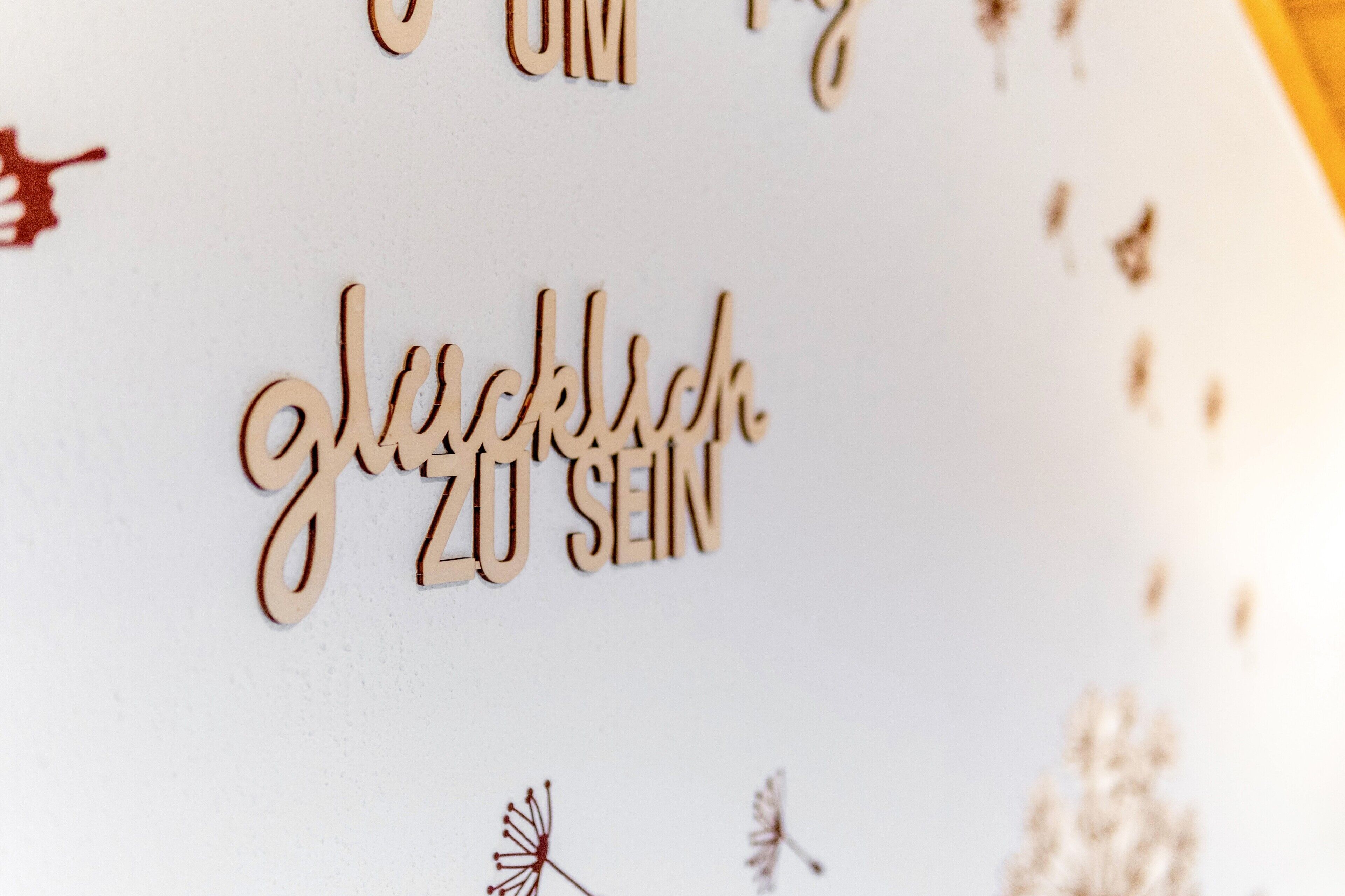 Une enseigne en bois portant l’inscription « glücklich zu sein », entourée de petites décorations florales stylisées.