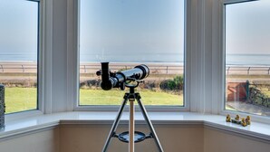 Een telescoop op een statief in een kamer met grote ramen met uitzicht op een strand.