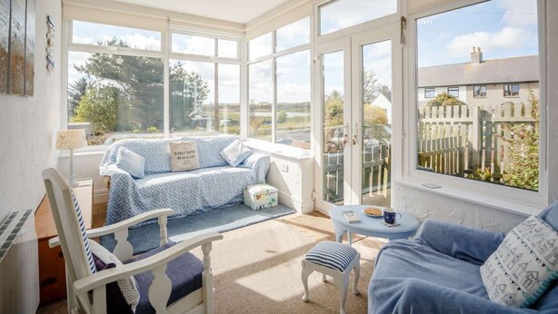 A bright room with large windows, a bed with a blue and white patterned bedspread, a white chair, a small round table with a cup and saucer, and a hanging light fixture. - 3 Bed in Low Newton By The Sea (oc-cn203) (Newton-by-the-Sea)