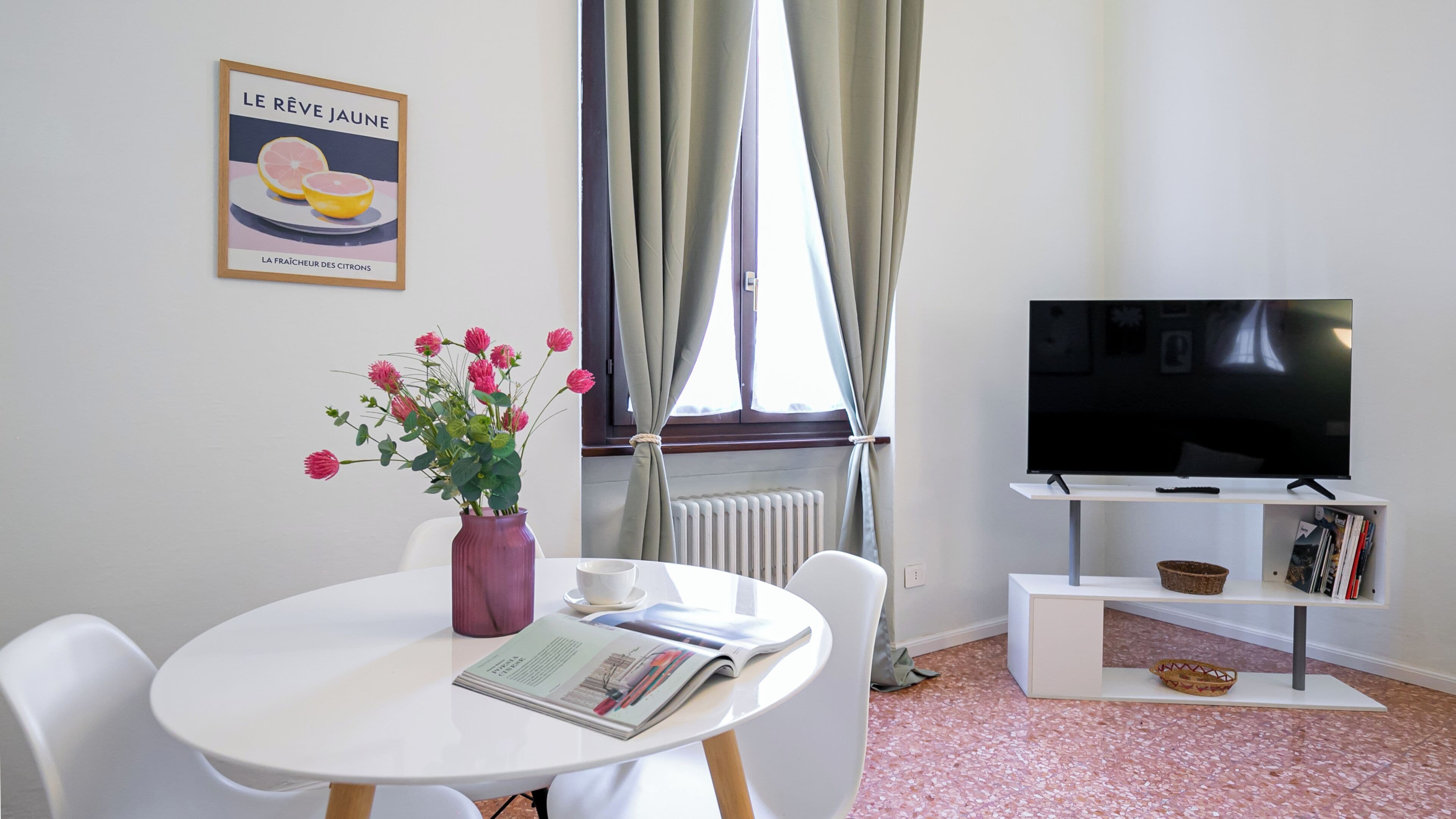 Une pièce comprenant une table à manger, un vase de fleurs, un tableau encadré accroché au mur, un radiateur, une fenêtre avec des rideaux, un téléviseur posé sur un meuble et une bibliothèque.