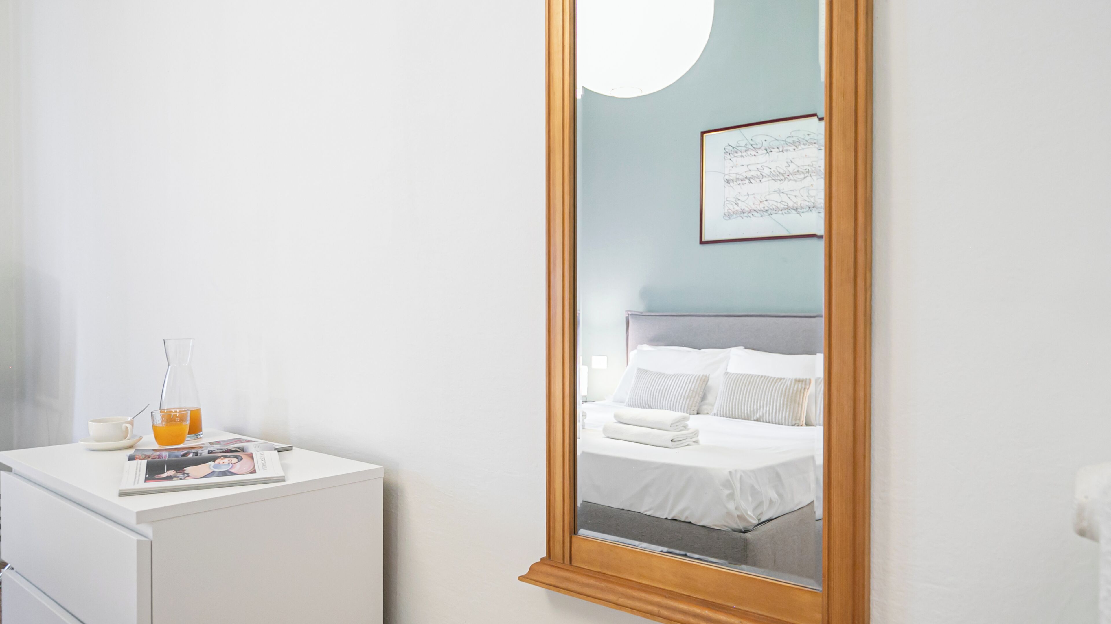 A hotel room with a bed, a bedside table with a magazine, a glass pitcher of orange juice, and a framed picture on the wall.