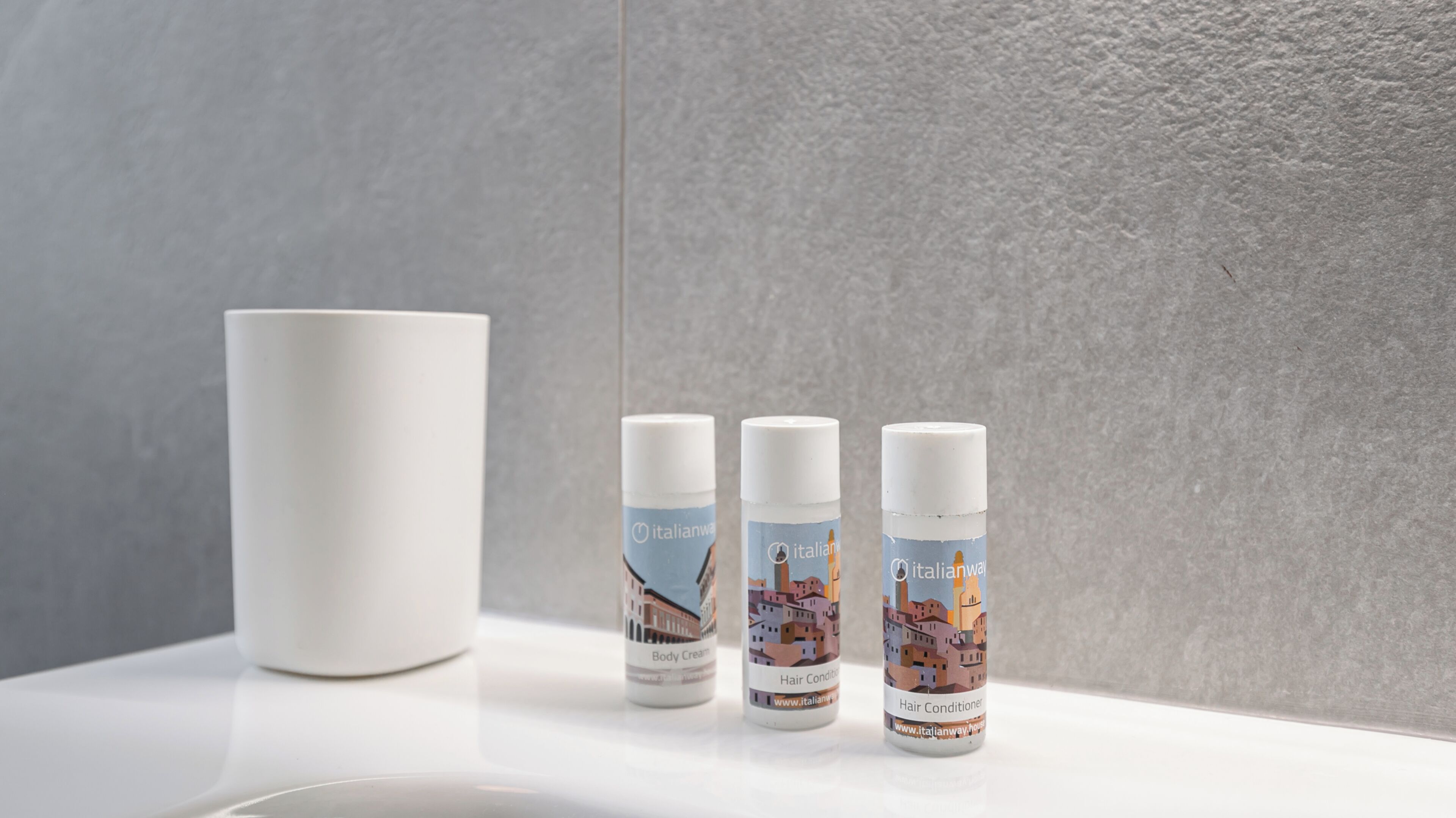 Trois flacons d’après-shampoing Italianway et une tasse blanche sur une surface blanche, le tout placé devant un mur gris.
